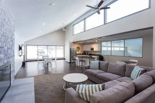 Open-concept living space with gray sectional sofa, kitchen, large windows, and high angled ceiling.