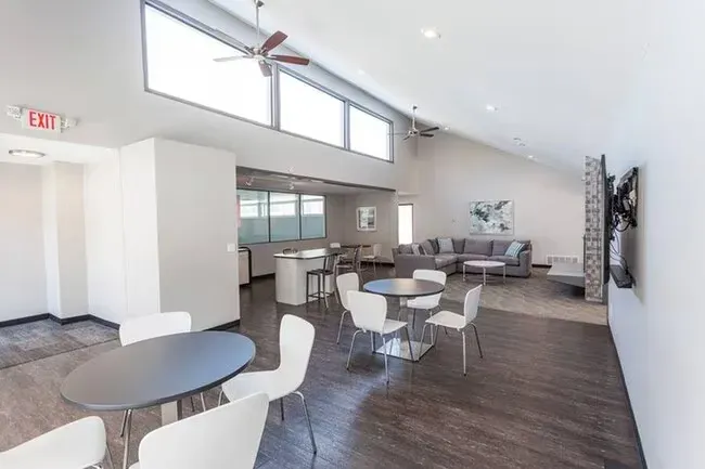 Modern lounge area with tables and chairs, sofa, and a kitchen area, with a TV on the wall.