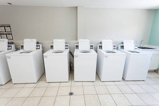 Row of white washing machines in a laundry room, tile floor, light walls.
