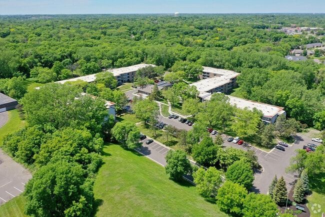 Aerial view of apartment complex surrounded by lush green trees and a park-like setting with parking.