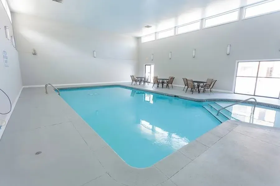 Indoor swimming pool with light blue water and grey walls. Tables and chairs near the pool's edge.