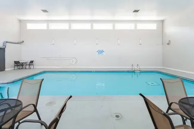 Indoor swimming pool with lounge chairs and tables. Light blue water, white walls, and bright overhead windows.