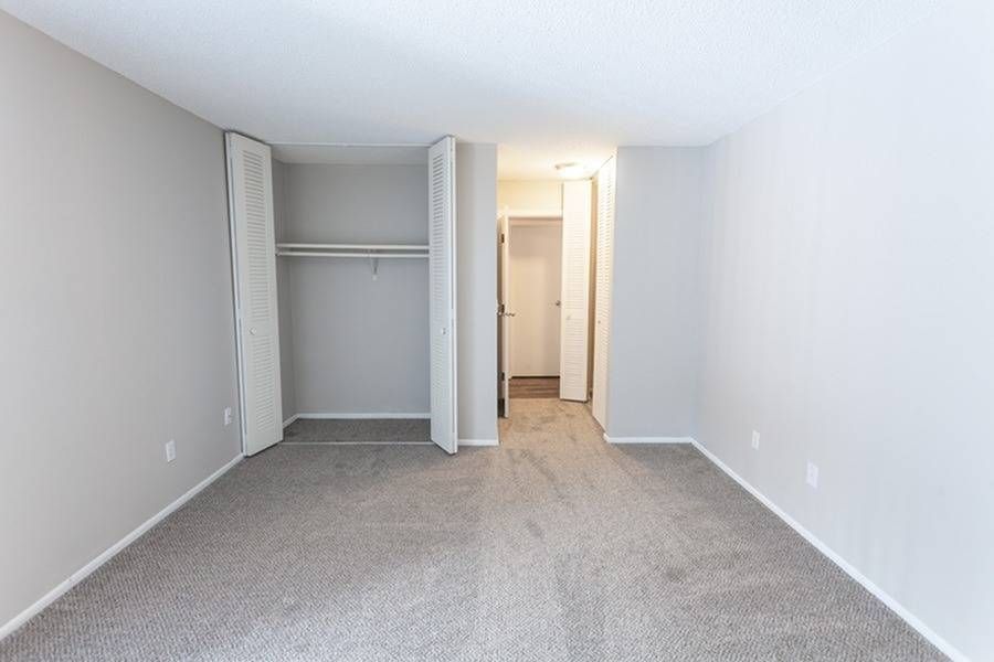 Empty apartment bedroom with an open closet, neutral walls, and carpet.