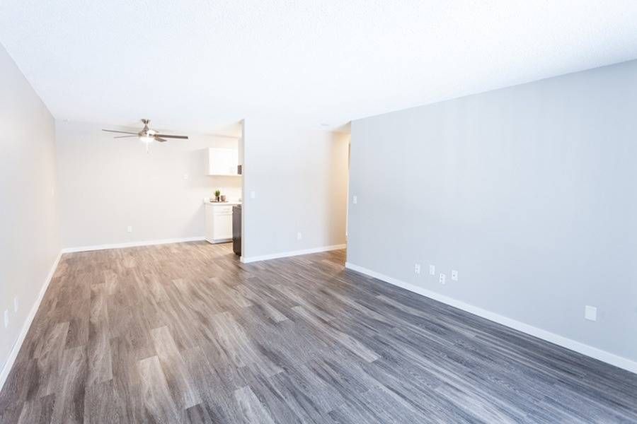 Empty living room with gray walls, wood-look flooring, and a small kitchenette in the back.