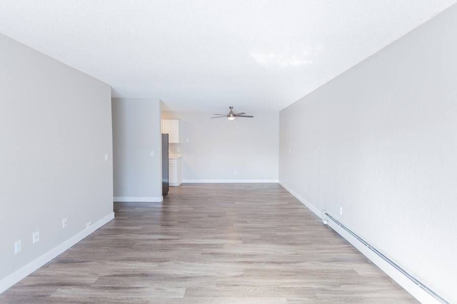 Empty living area with light gray walls, wood-look flooring, baseboard heating, and a ceiling fan.