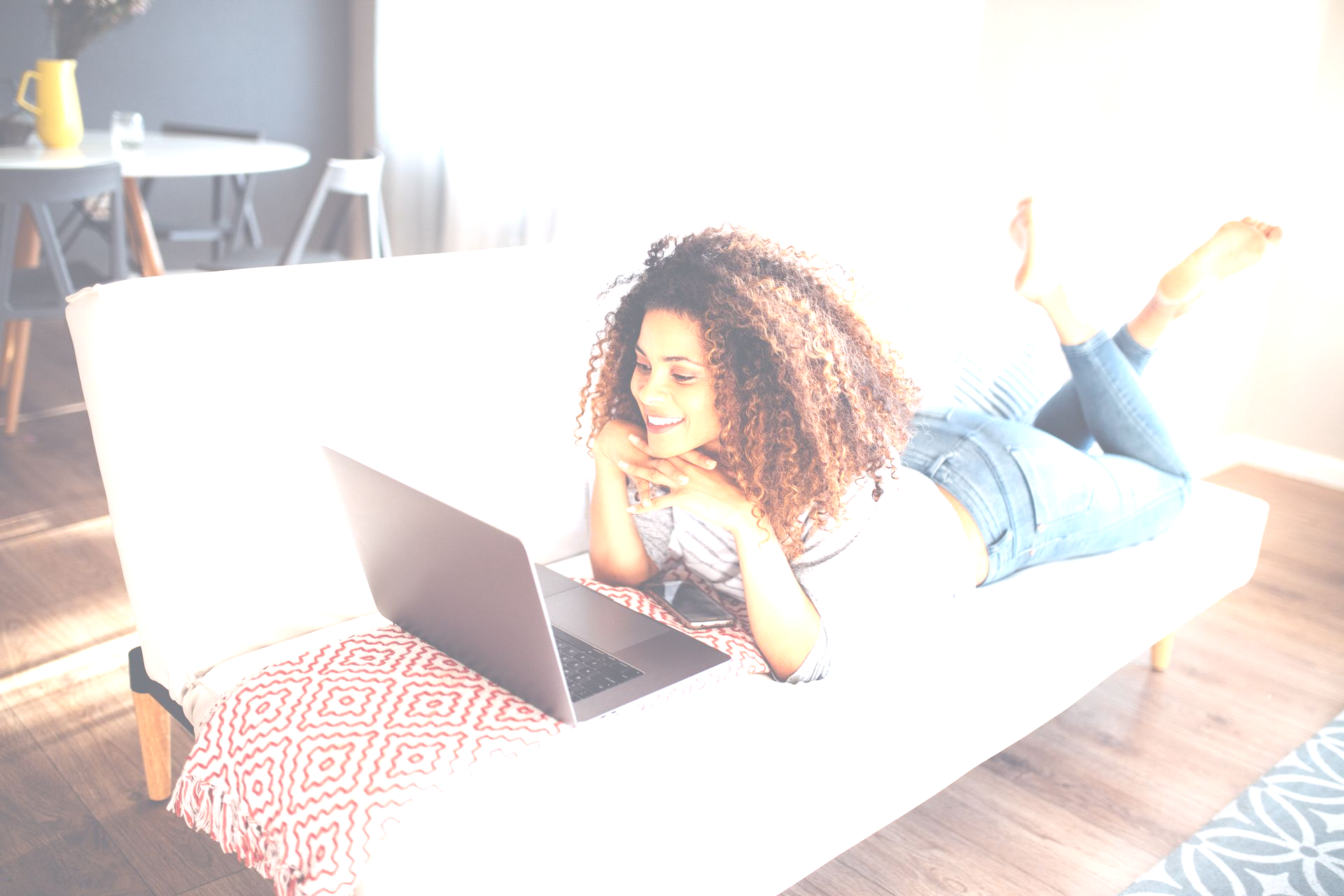 Woman with curly hair, smiling while using a laptop on a white couch in a bright living room at Talus Apartment Homes