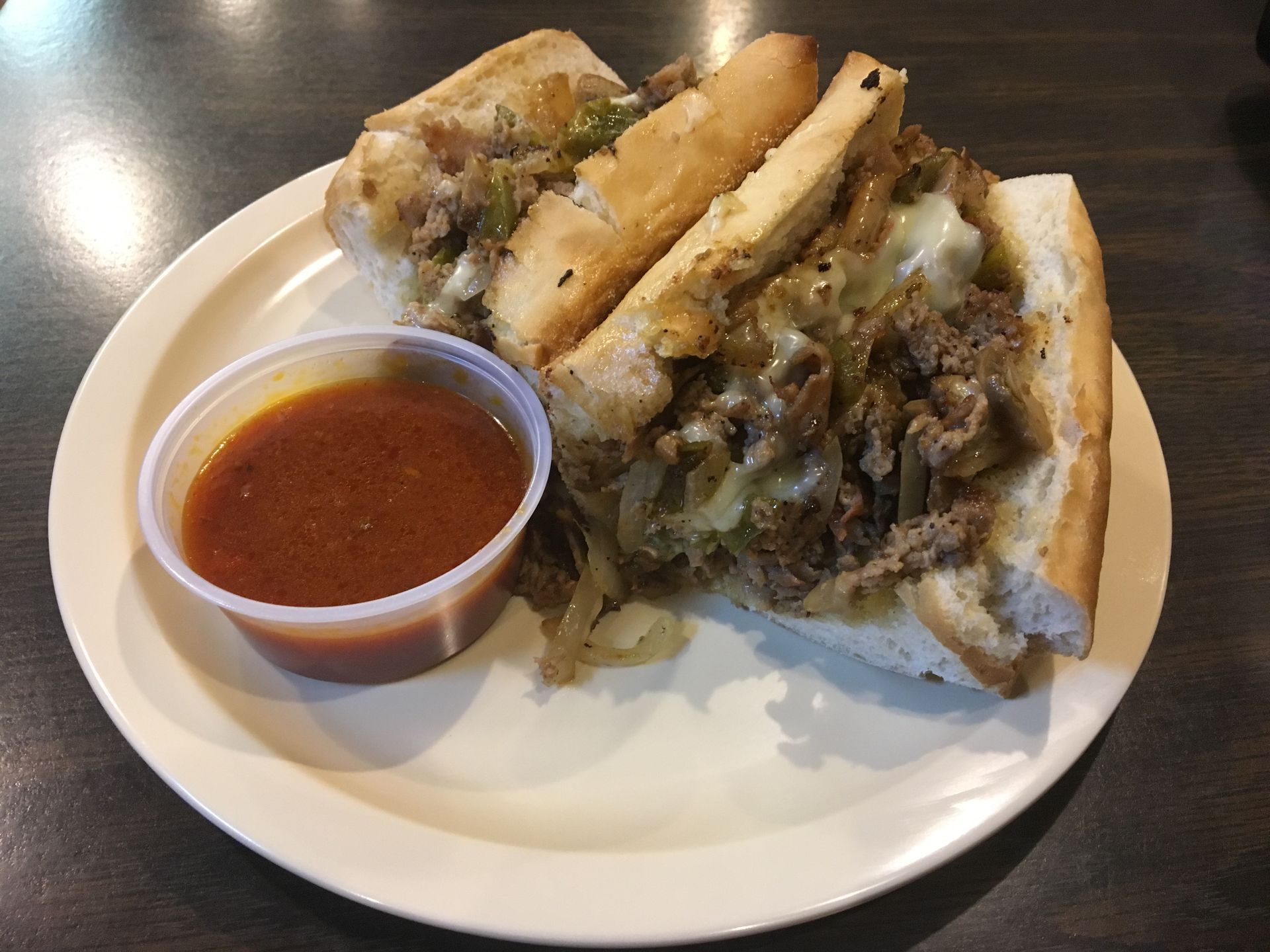 A steak and cheese sub sandwich with a side of sauce on a white plate.