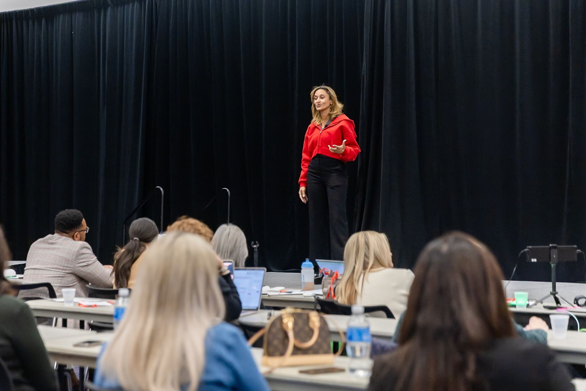 Speaker in red blouse addressing an audience in a conference room with black curtains.