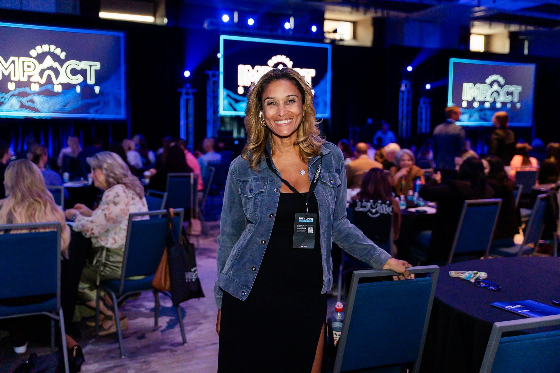Smiling woman in a blue blazer stands at a conference event with screens and seated attendees in the background