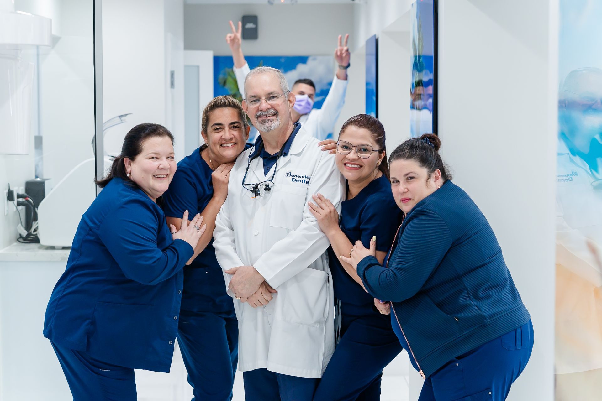 Dental staff pose together in a brightly lit office; smiling, wearing blue scrubs, doctor in lab coat.