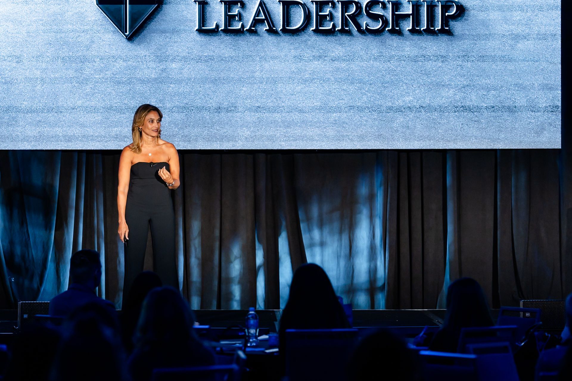 Speaker on stage in black dress, standing before a “Leadership” backdrop and audience silhouettes