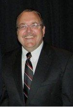 Man in suit and tie smiling, wearing glasses. Black background.