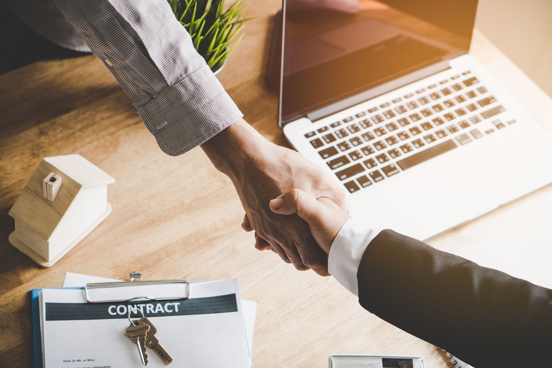 Two people shaking hands over a contract and keys. Laptop and house model on desk.