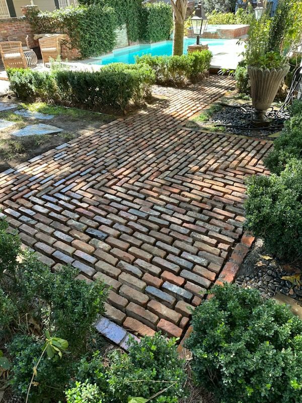 A brick walkway in a garden with a pool in the background.