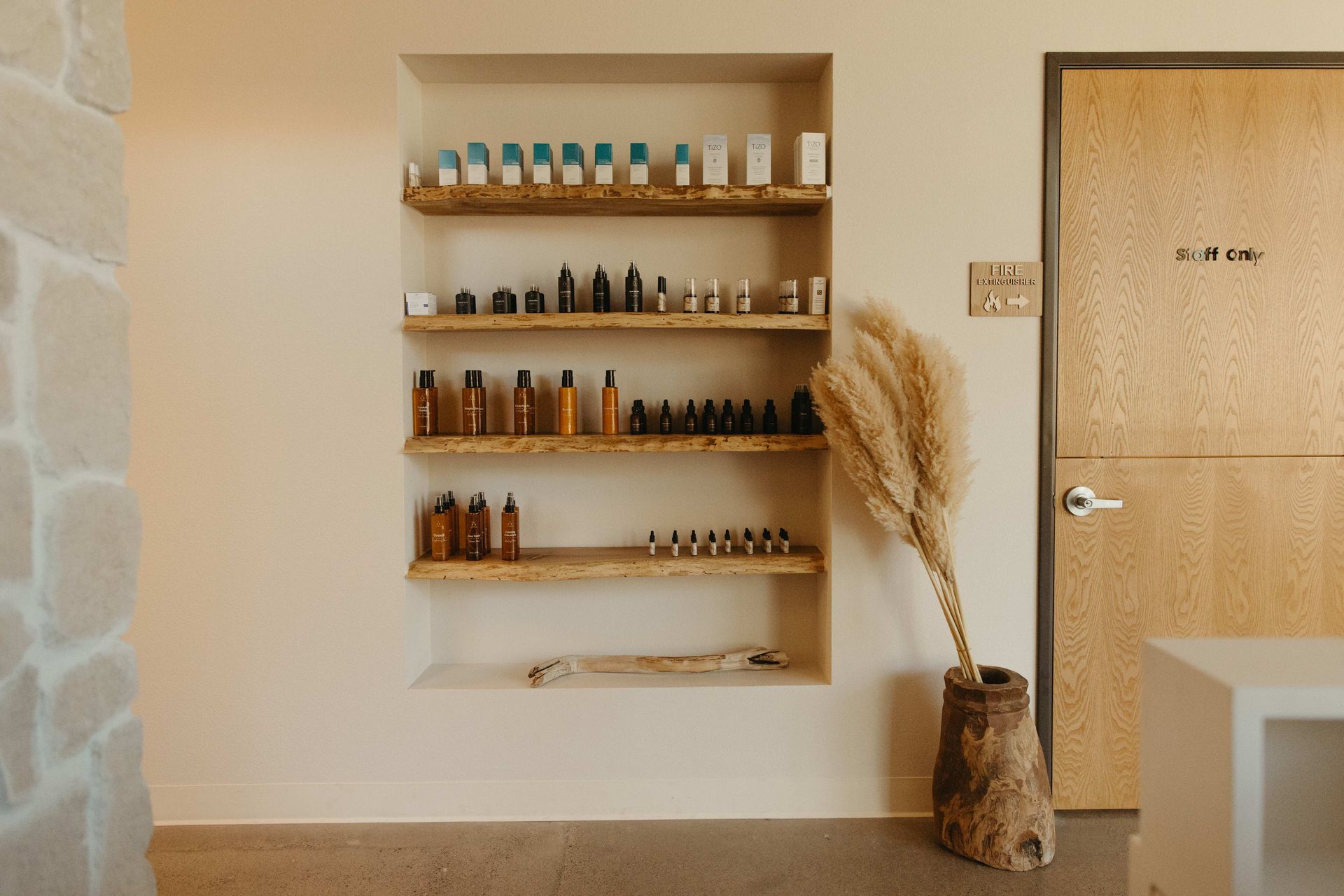 Shelves with cosmetic products in a recessed wall next to a door and dried plant in a rustic vase.