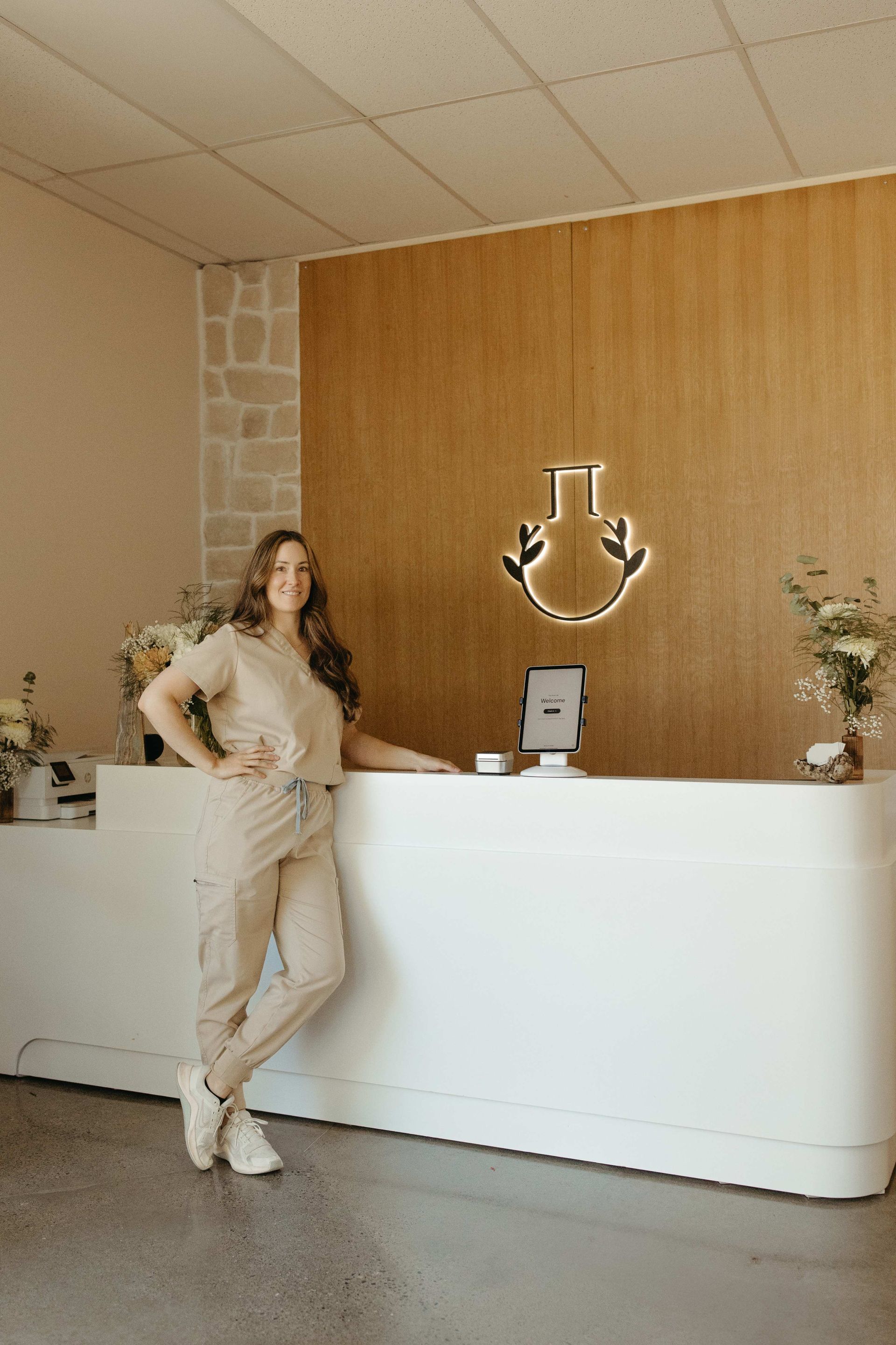 Woman in tan scrubs stands at a white reception desk, smiling in a modern office with gold accents.