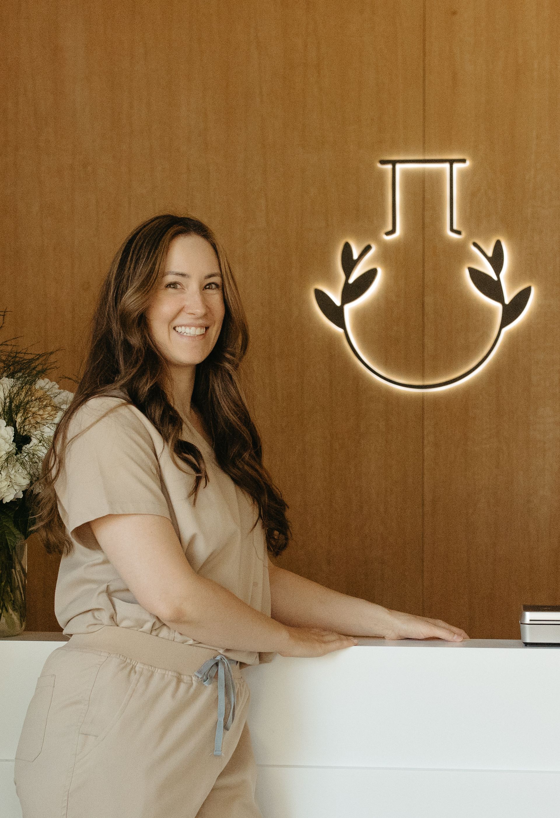 Woman in tan scrubs smiles, stands behind a white counter, with a gold logo on a wood wall.