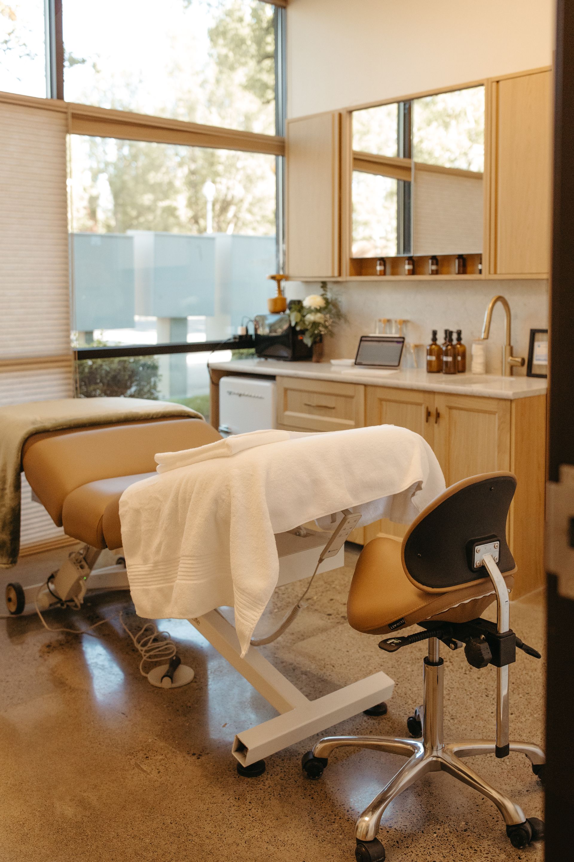 Massage therapy room with a tan table, white towels, and a rolling chair. Light wood cabinets and large window.