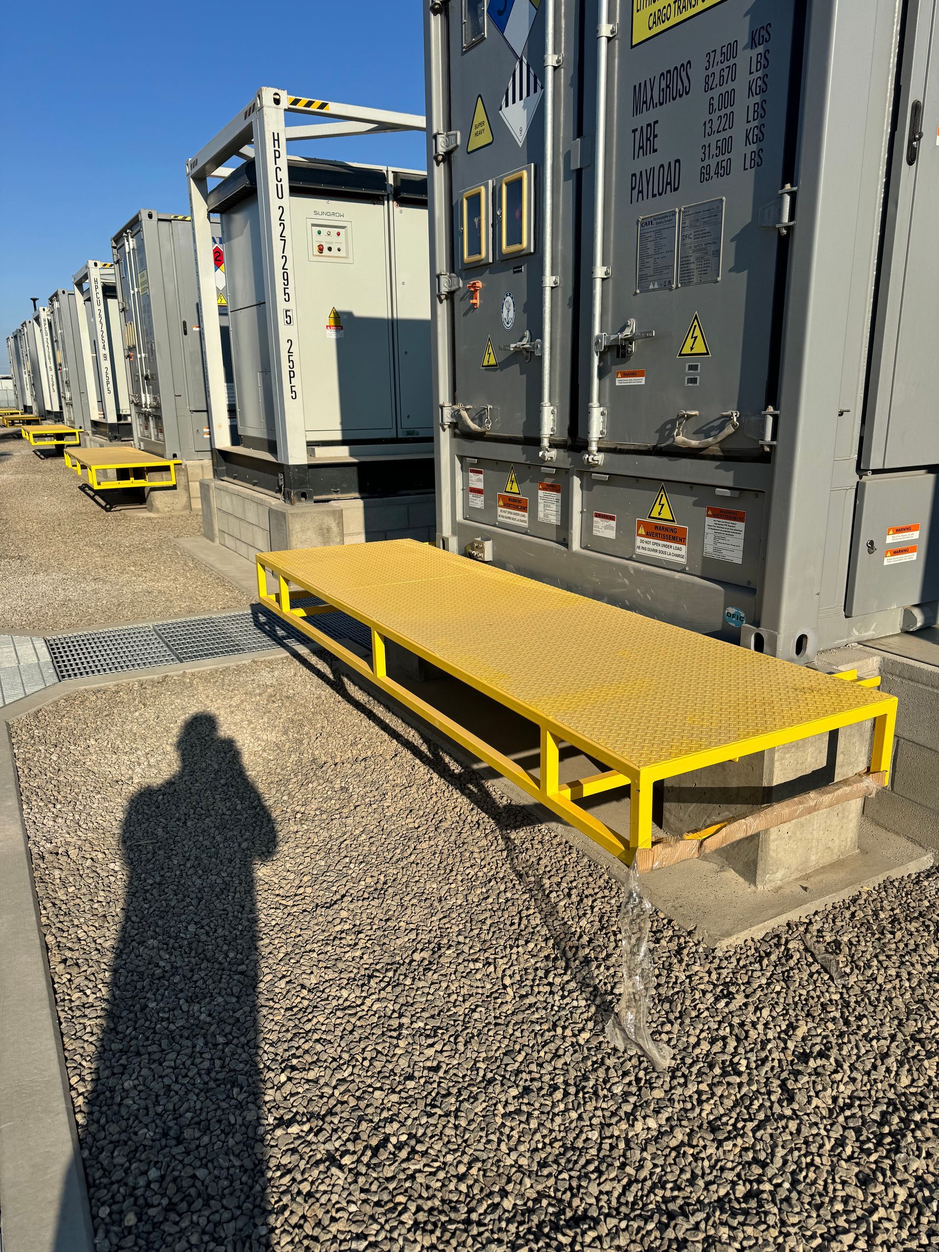 Yellow metal ramp platform in front of a gray industrial container, on gravel ground. Shadow of a person present.
