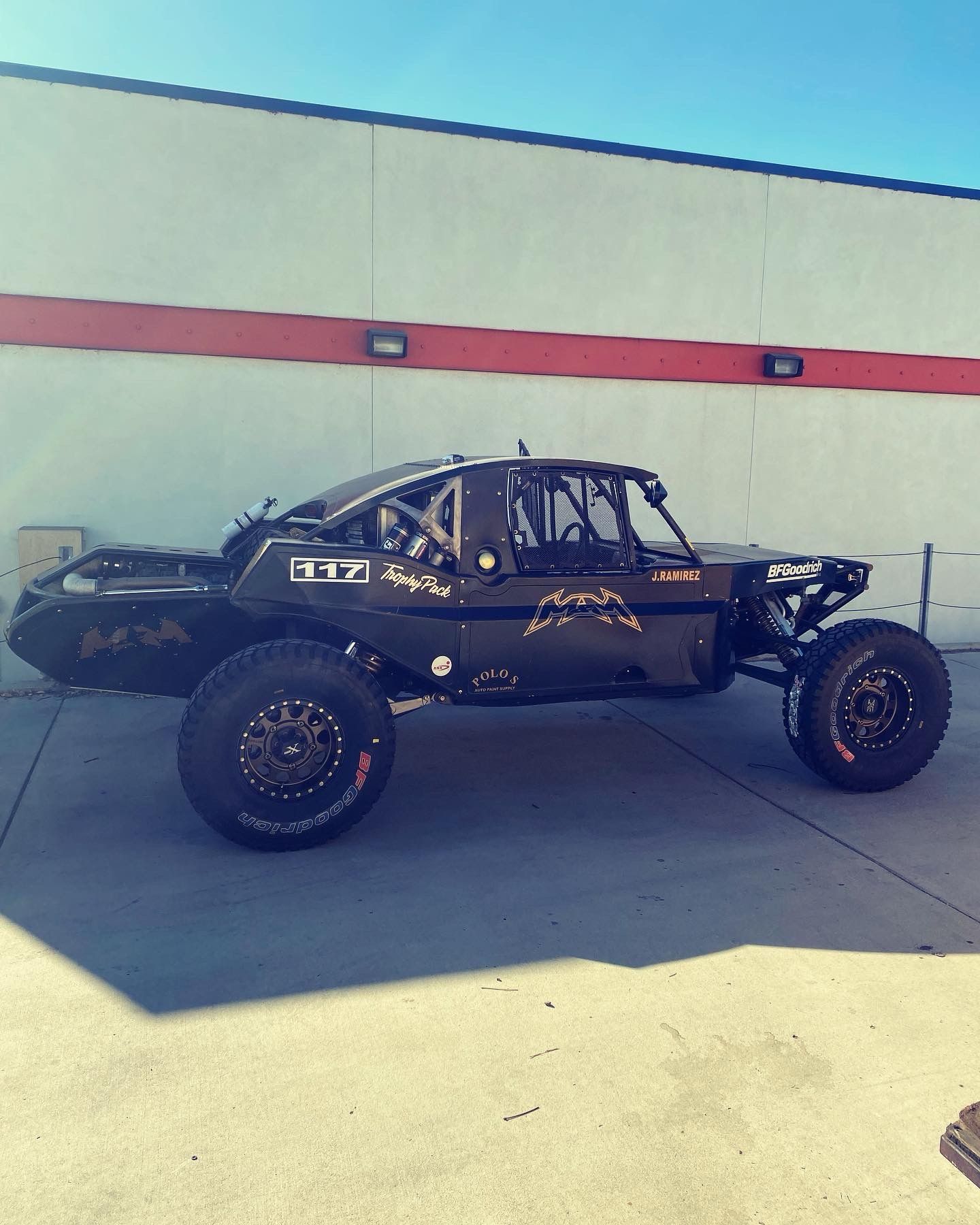 Black off-road race truck parked in front of a white building with red stripe.