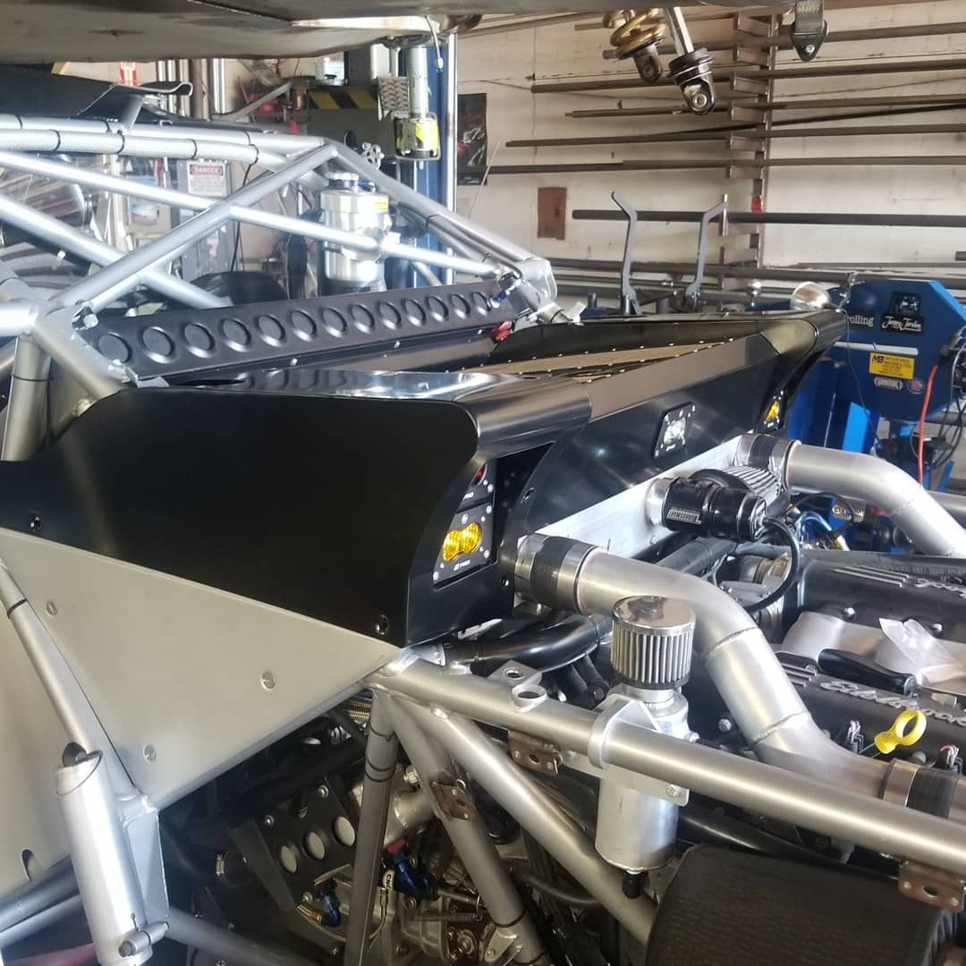 Race car chassis in a workshop, exposed engine, roll cage. Silver, black, and mechanical details visible.