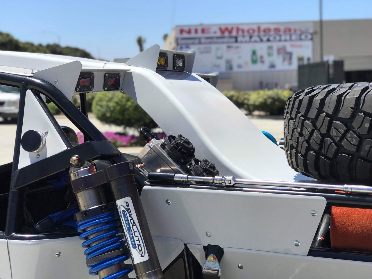Off-road vehicle rear details: white frame, spare tire, shock absorber, lights, and a building with a sign in the background.