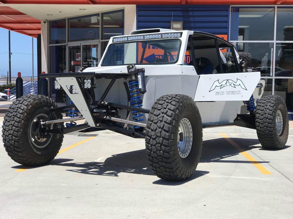 White off-road race buggy with large tires, blue shocks, parked outside a building.