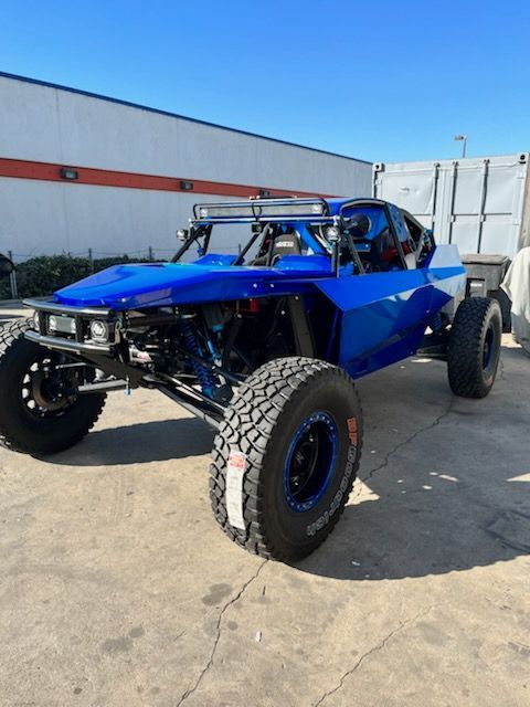 Blue off-road racing vehicle with large tires, roll cage, and light bar, parked outdoors.