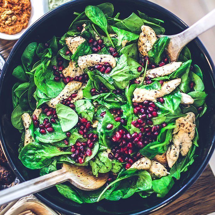 A salad with spinach , chicken , pomegranate seeds and a wooden spoon in a bowl.