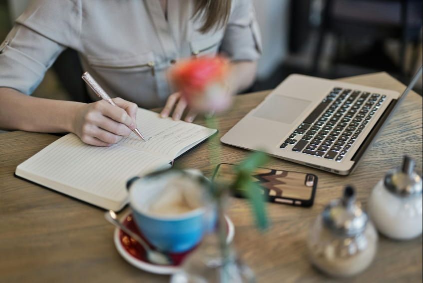A woman is writing in a notebook while sitting at a table with a laptop and a cup of coffee.