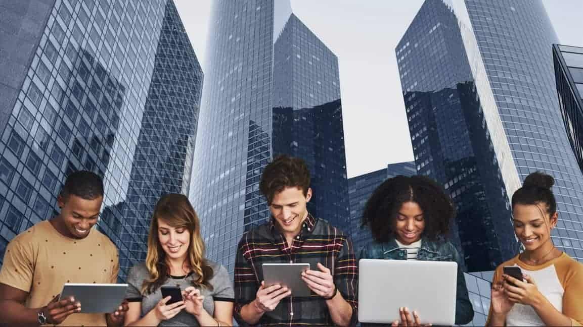A group of people are standing next to each other looking at their phones and laptops.