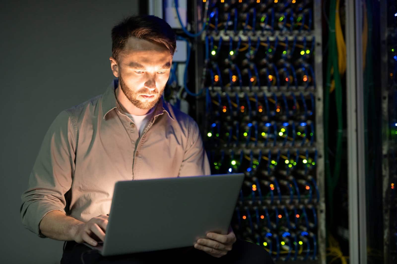 A man is using a laptop computer in a server room.