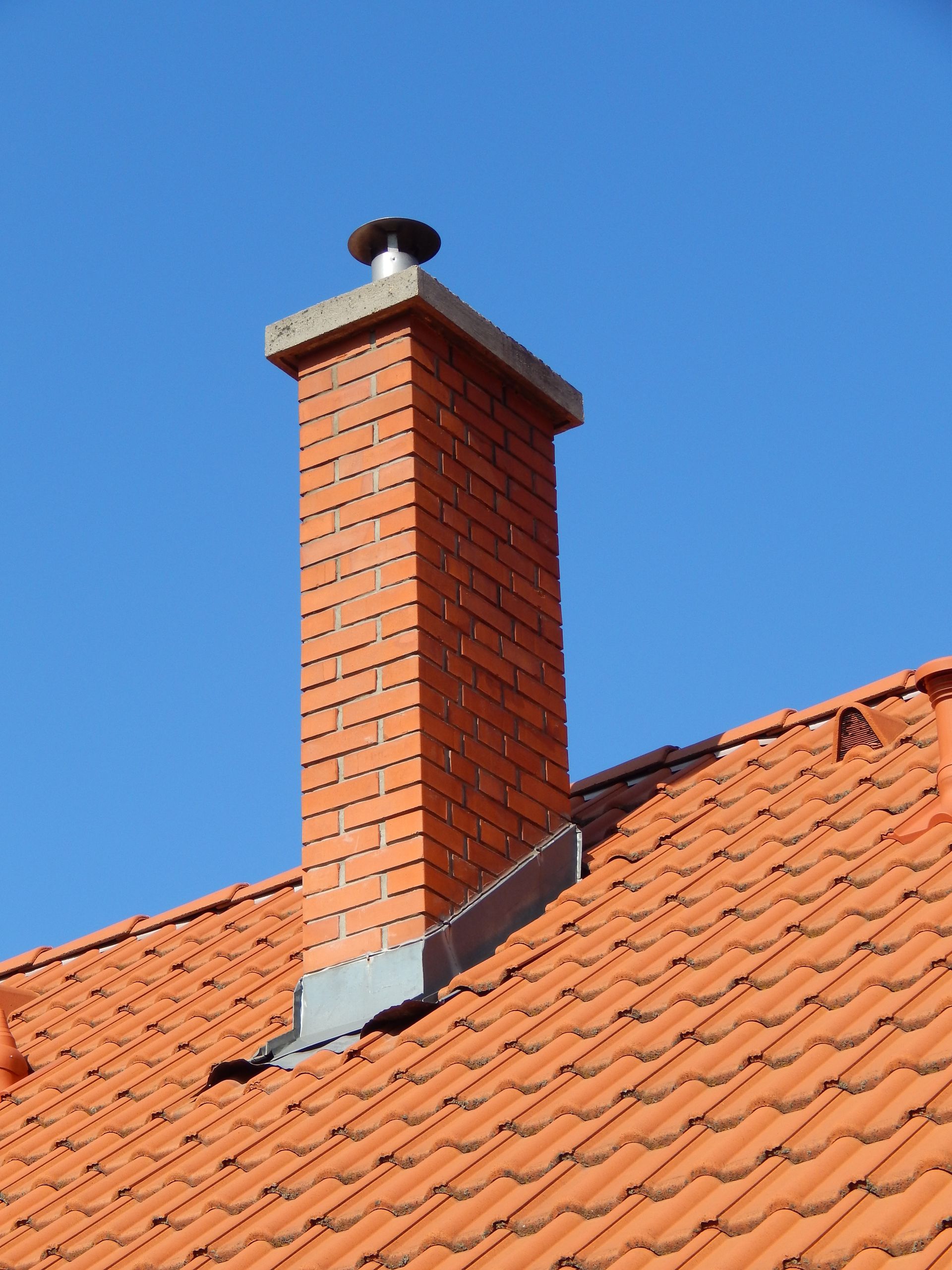 Red brick chimney on a terracotta tile roof against a clear blue sky.