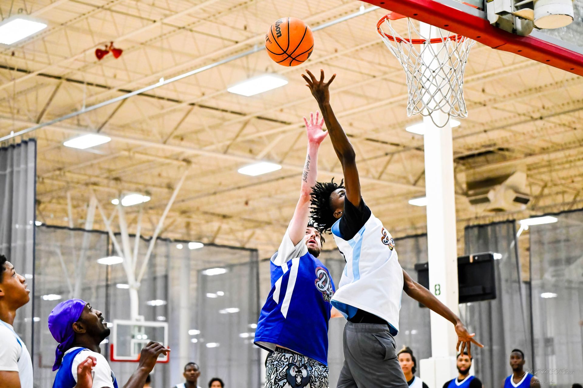 Two basketball players jump toward a hoop in an indoor gym, one in a white jersey and the other in blue, competing for ball.