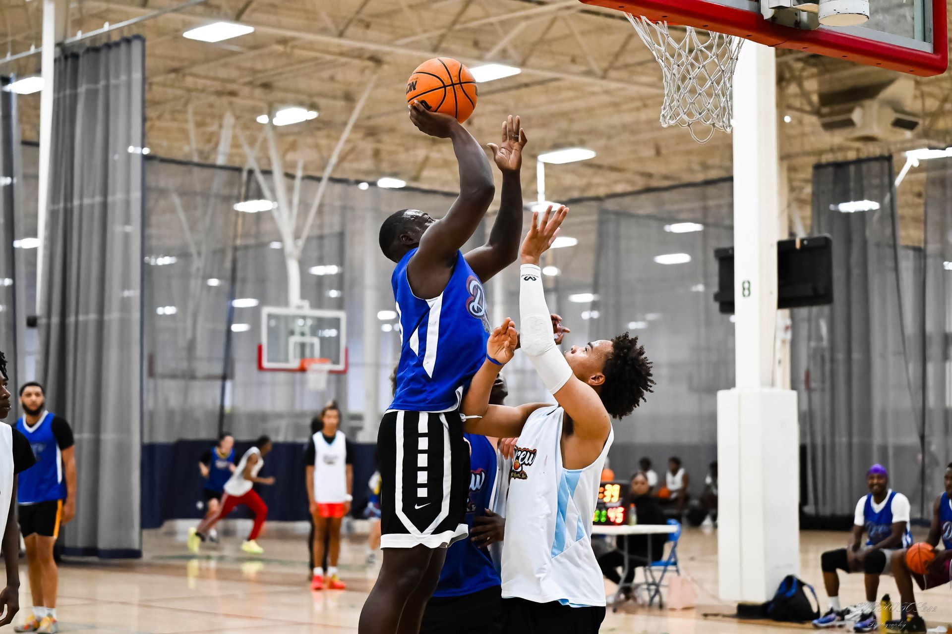 Two basketball players in an indoor arena jump toward the hoop to contest a shot, wearing blue and white jerseys.