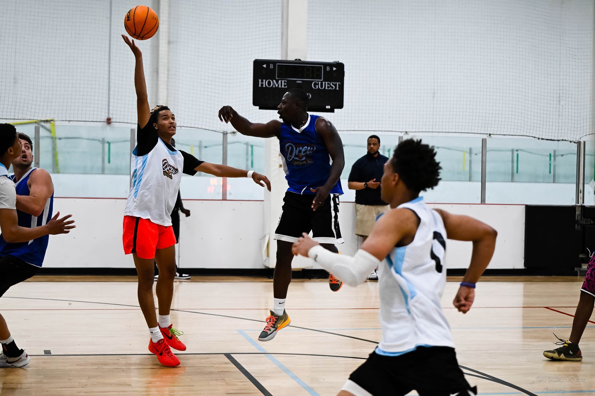 Athletes play basketball on an indoor court, with one player jumping to reach for the ball while others watch.