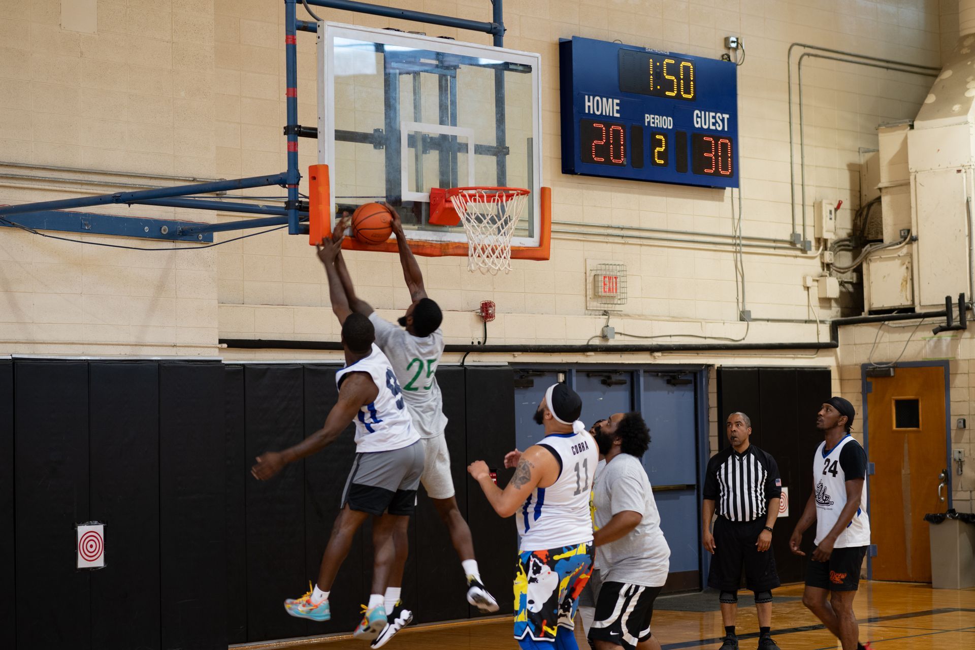 Two basketball players jump to contest a shot at the hoop while teammates and a referee watch in a gymnasium.