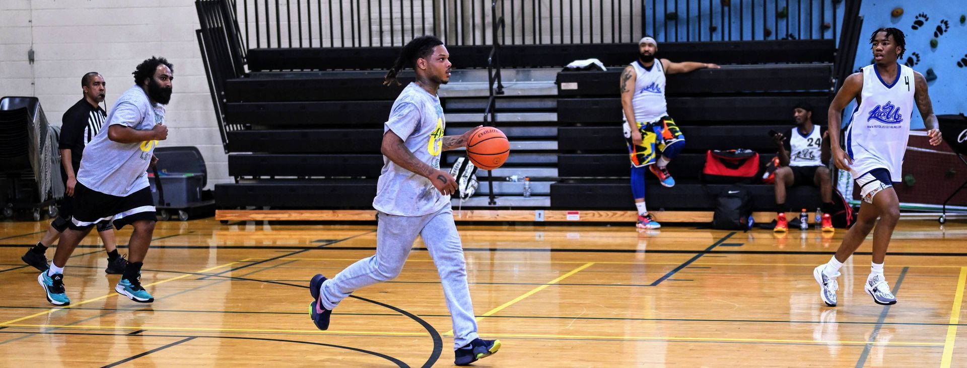 Players in athletic wear practice basketball on an indoor court with wooden floors and bleachers in the background.