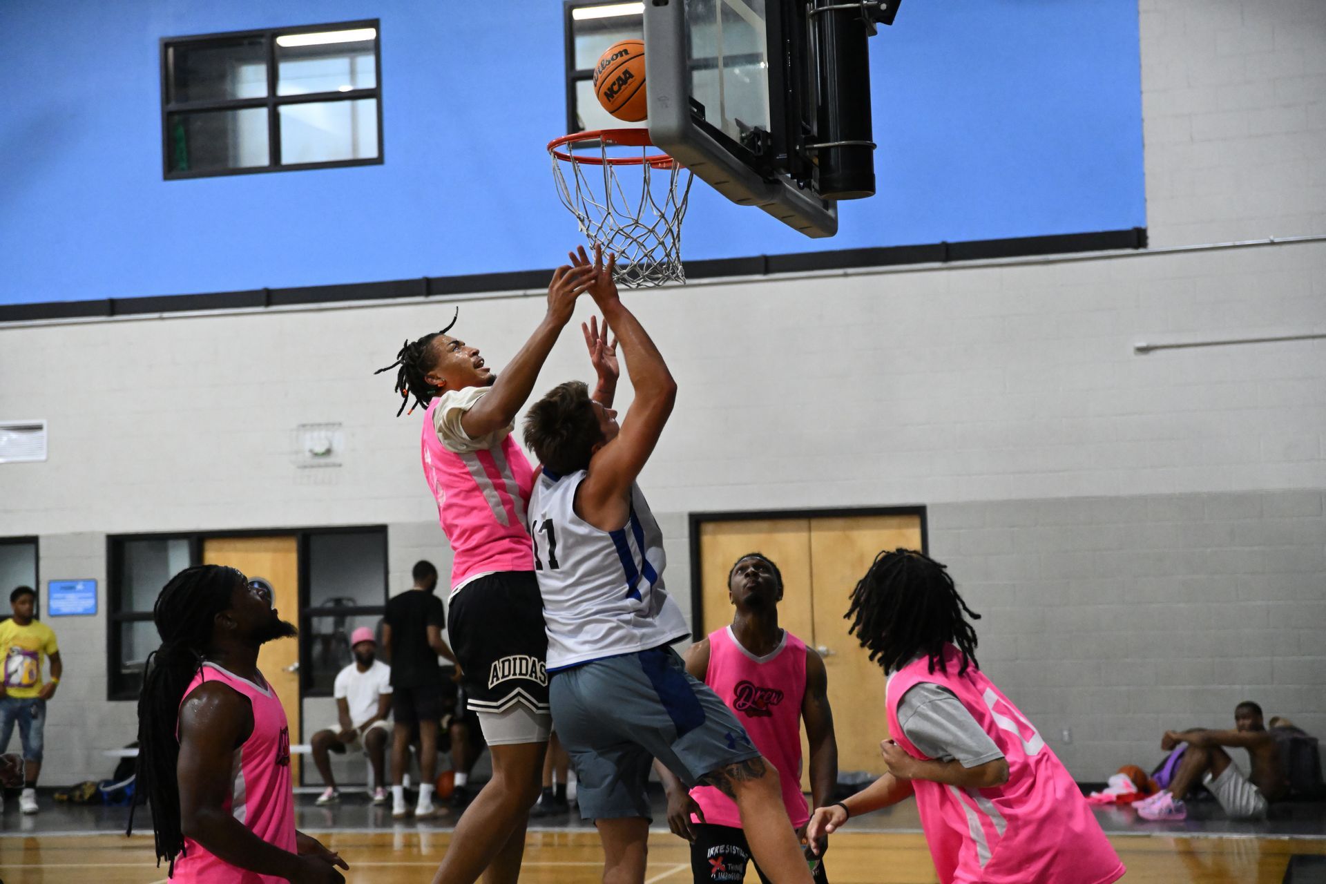 Two players jump to shoot a basketball into a hoop in an indoor gym, surrounded by other players in pink jerseys.
