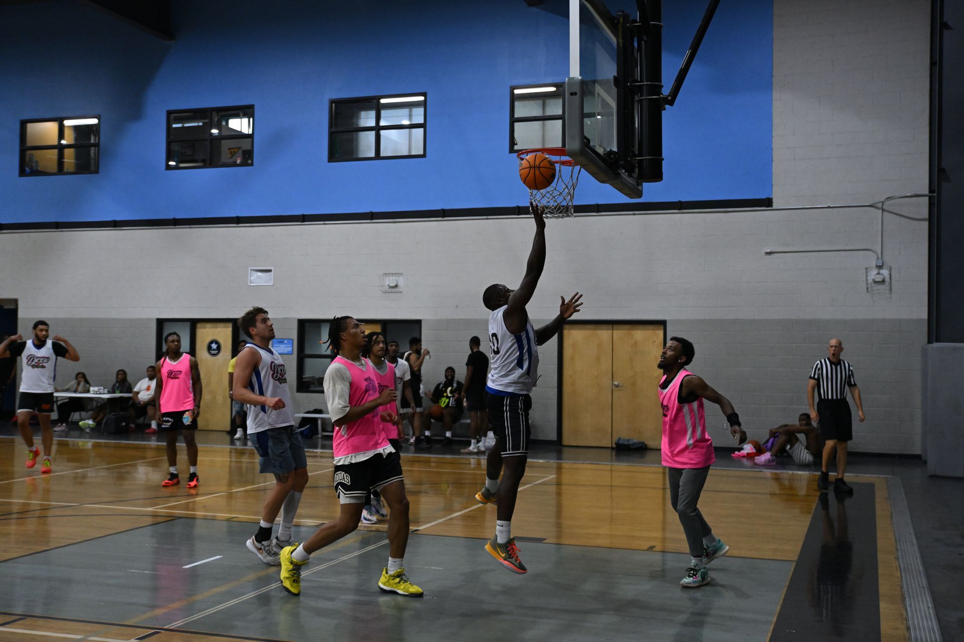 A basketball player in a white uniform jumps to shoot a ball into a hoop at an indoor court with other players nearby.