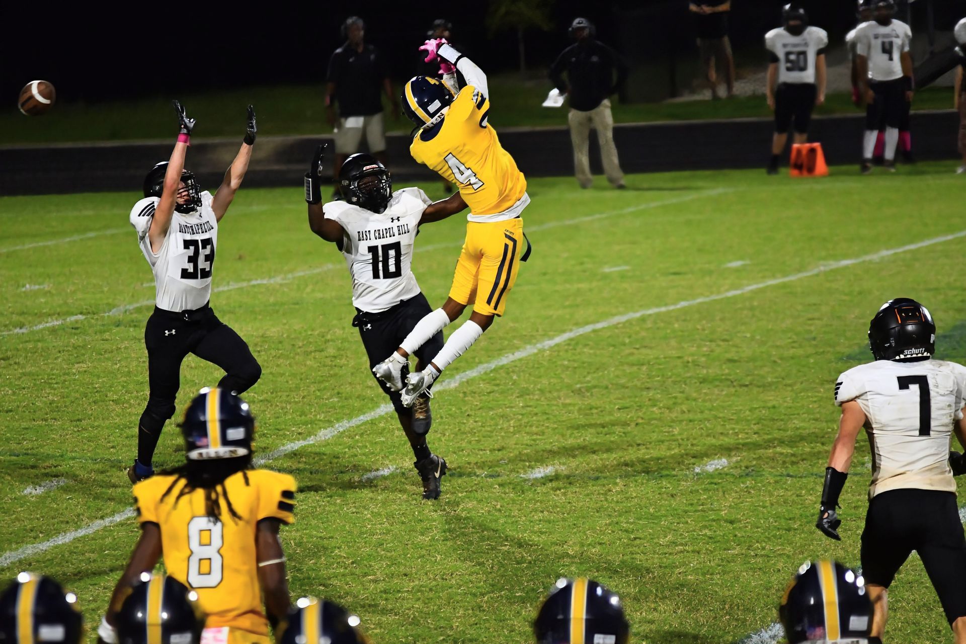 A yellow-jerseyed player jumps high to catch a football as two white-jerseyed defenders reach toward them on a field.