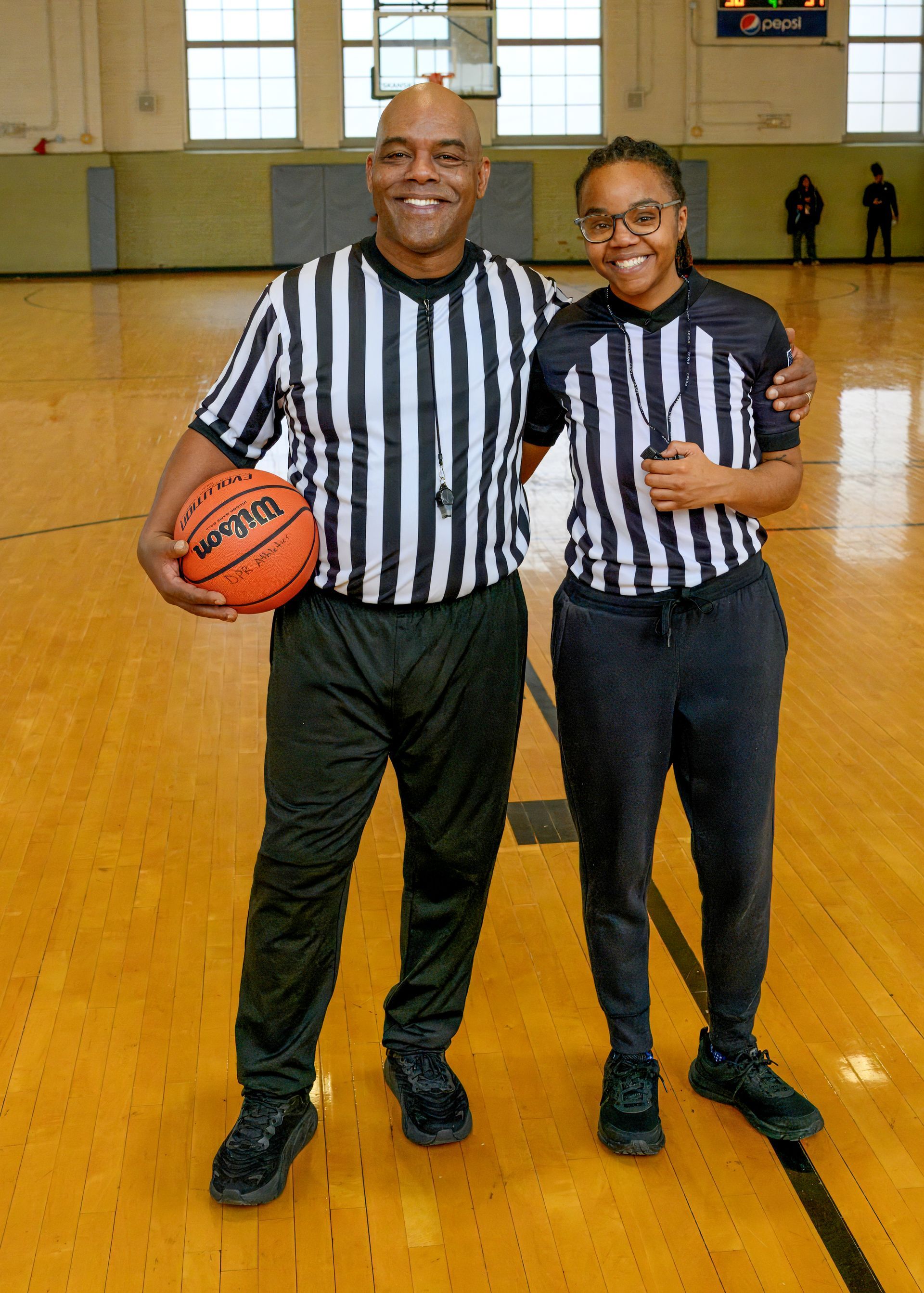 Two basketball referees stand on a court, one holding a basketball and both wearing black and white striped shirts.
