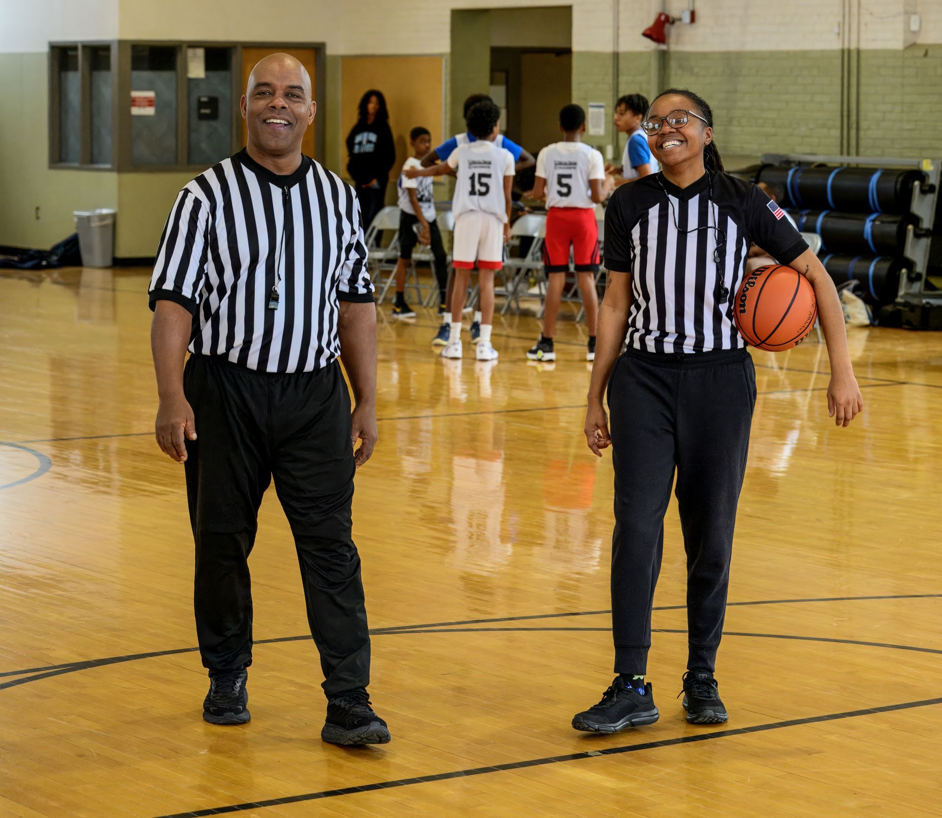 Two basketball referees wearing black-and-white striped shirts stand smiling on a court, one holding a basketball.