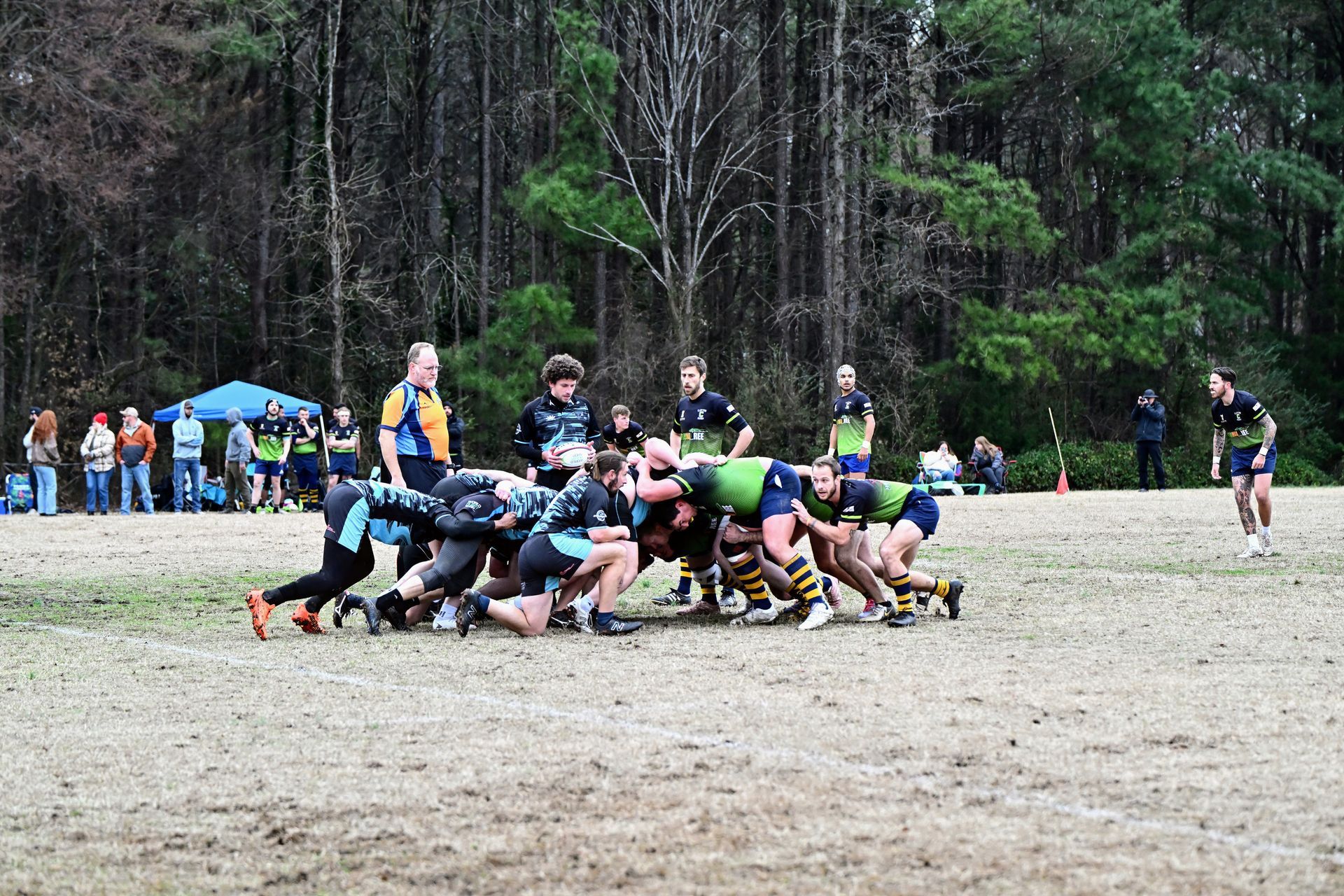 Rugby players in green and black uniforms engage in a scrum on a field in front of a tree line.