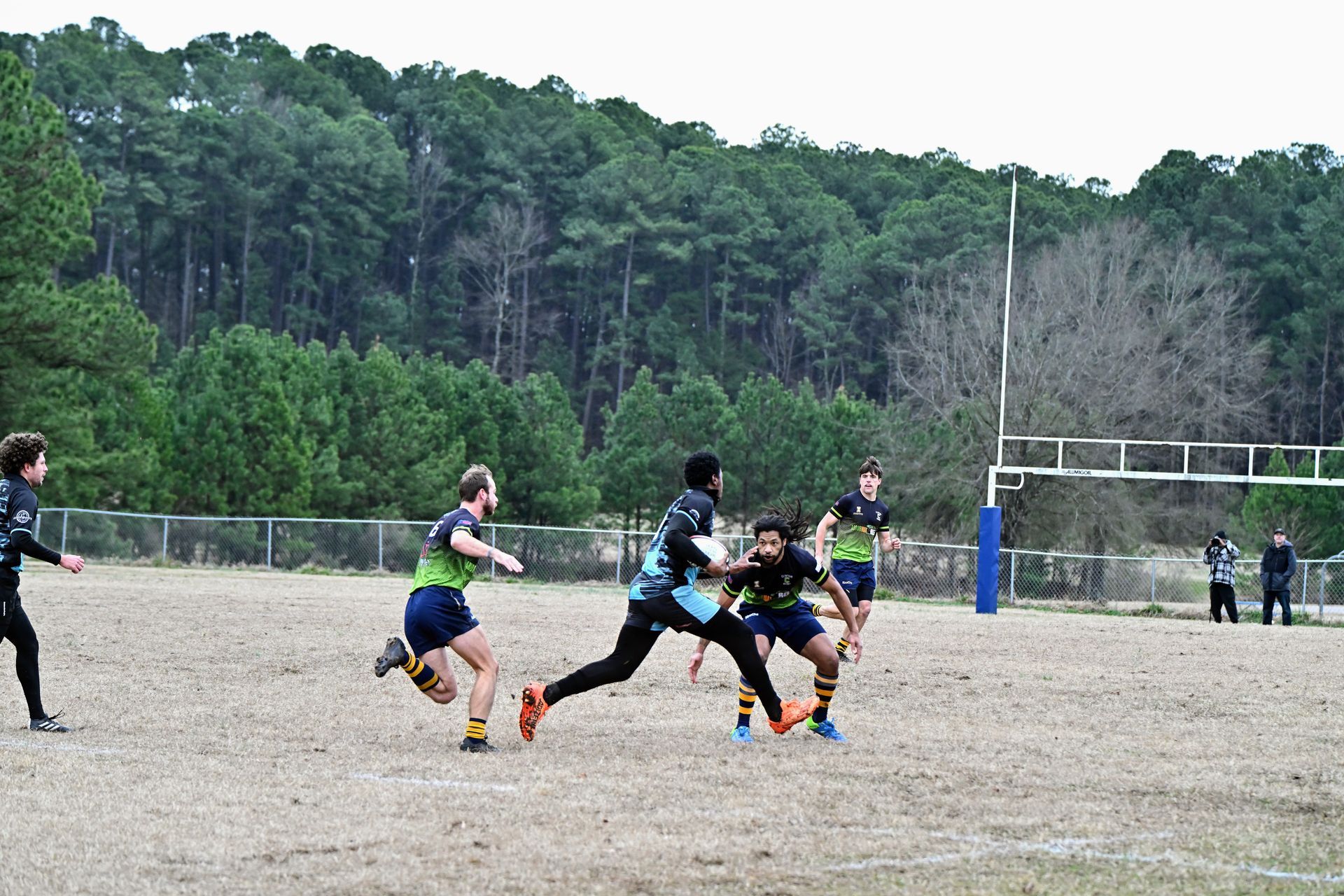 Players in green and blue rugby uniforms compete on a dirt field with a forest in the background.