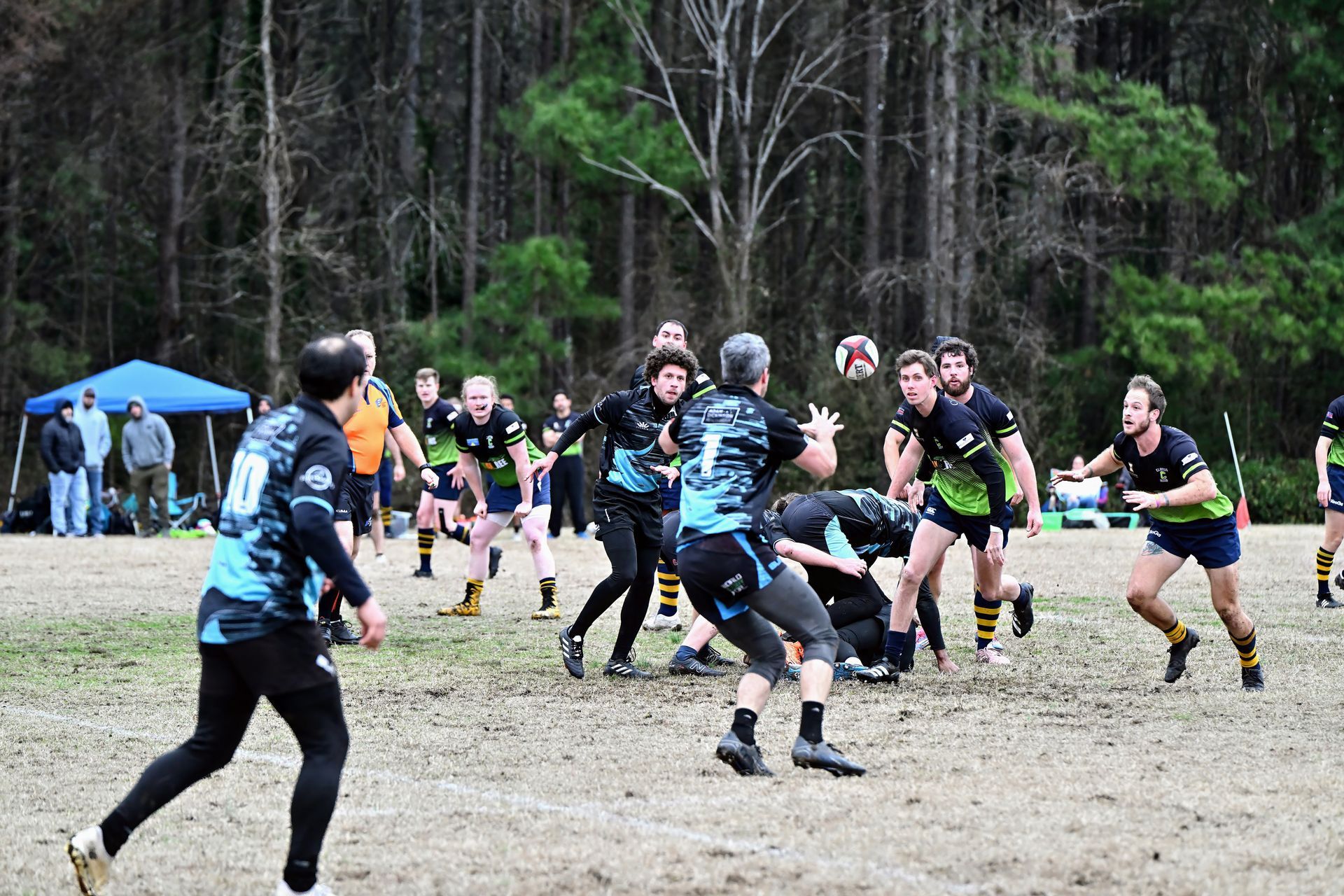 Rugby players in blue and black or green and black uniforms compete for a ball on a dirt field with trees in the background.