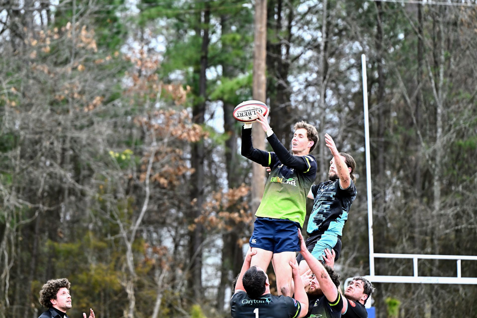 Rugby players compete for a ball mid-air, lifted by teammates in a lineout on a field with trees in the background.