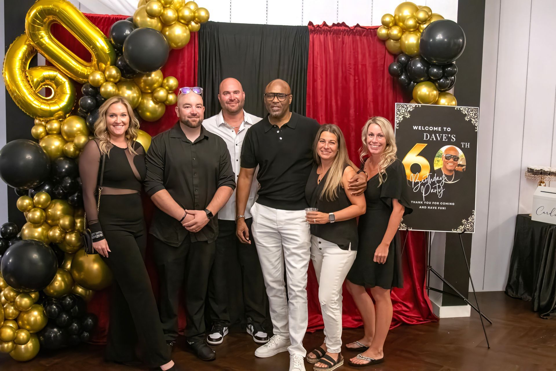 Five adults pose for a photo in front of a red and black curtain decorated with gold and black balloons for a 60th party.