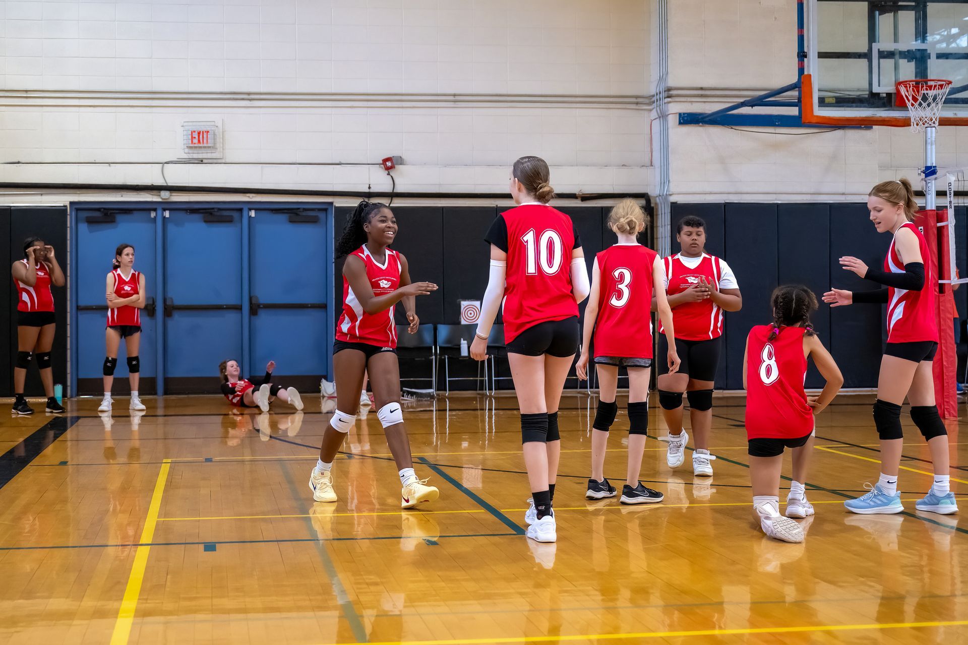Volleyball players in red uniforms celebrate on a hardwood court while one player sits on the floor in the background.