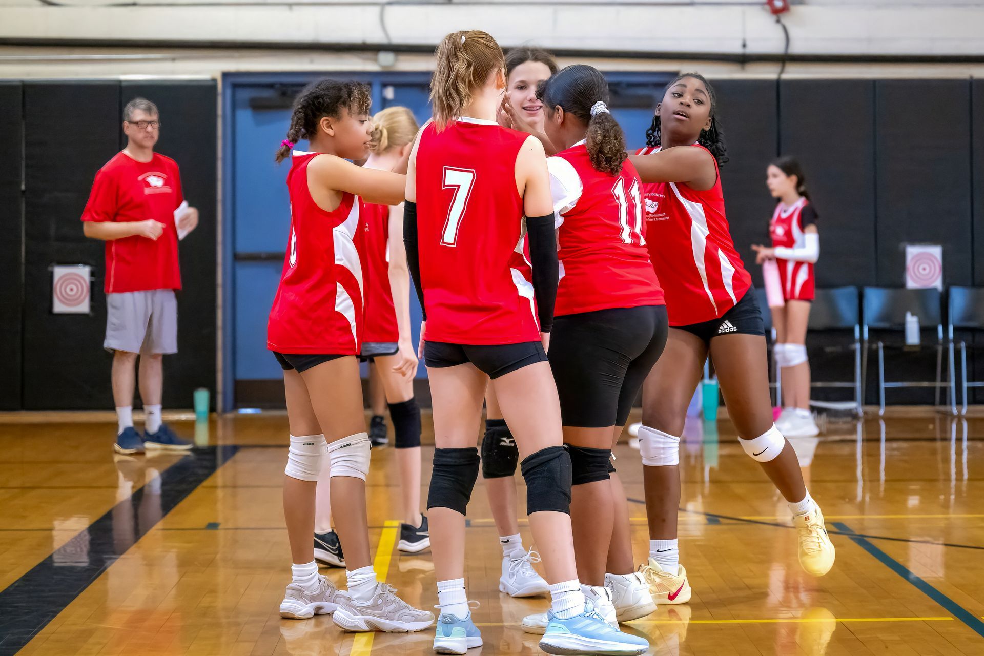 A group of volleyball players in red jerseys huddle on an indoor court, with a coach standing nearby in the background.